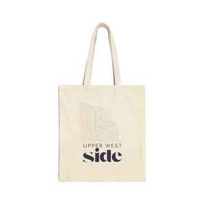 May include: Cream-colored tote bag with a minimalist design. The bag features a graphic of curved lines and the words "UPPER WEST SIDE" and "side" in navy blue. The tote has two handles for carrying.