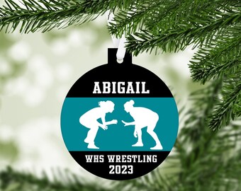 May include: A black and teal round Christmas ornament with a white silhouette of two wrestlers. The ornament has the name "Abigail" and the text "WHS Wrestling 2023".