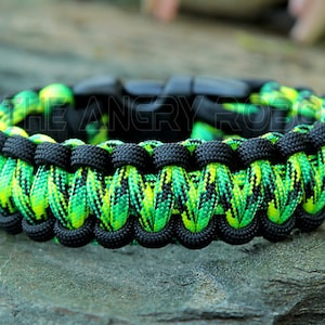 Puede incluir: Una pulsera de paracord negra y verde ne&oacute;n con un cierre negro. La pulsera est&aacute; hecha de cord&oacute;n de nailon tejido en un patr&oacute;n vibrante. El texto "THE ANGRY ROBOT" es visible en la pulsera. La pulsera est&aacute; sobre una superficie de piedra gris.