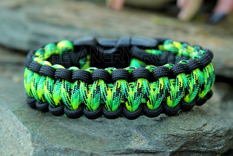 Puede incluir: Una pulsera de paracord negra y verde ne&oacute;n con un cierre negro. La pulsera est&aacute; hecha de cord&oacute;n de nailon tejido en un patr&oacute;n vibrante. El texto "THE ANGRY ROBOT" es visible en la pulsera. La pulsera est&aacute; sobre una superficie de piedra gris.