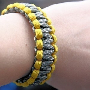 Puede incluir: Una pulsera trenzada en tonos grises y amarillos. La pulsera est&aacute; hecha de cord&oacute;n tejido, con el cord&oacute;n amarillo formando un patr&oacute;n alrededor del gris. La pulsera mide aproximadamente 2 cm de ancho.