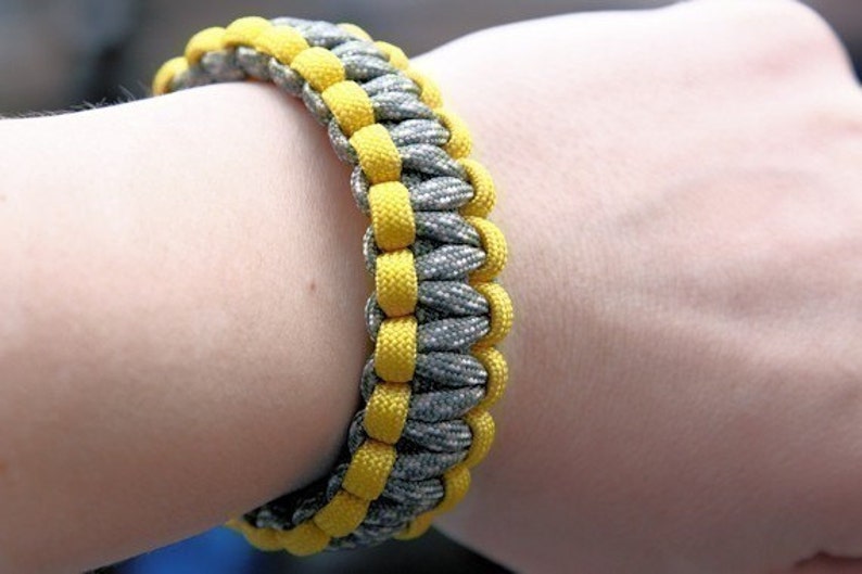 Puede incluir: Una pulsera trenzada en tonos grises y amarillos. La pulsera est&aacute; hecha de cord&oacute;n tejido, con el cord&oacute;n amarillo formando un patr&oacute;n alrededor del gris. La pulsera mide aproximadamente 2 cm de ancho.