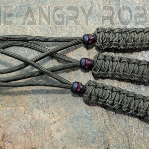 May include: Three green paracord keychains with black skull beads and black plastic tips. The keychains are on a gray rock surface.