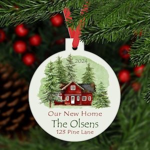 May include: A white round Christmas ornament with a watercolor illustration of a red house with green trees and the text "Our New Home The Olsens 123 Pine Lane 2024".