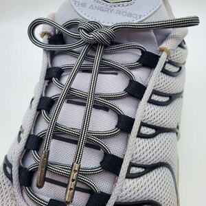 May include: Close-up of a pair of white sneakers with black and white striped shoelaces tied in a knot. The laces are threaded through the eyelets and the text "THE ANGRY ROBOT" is visible on the shoe.