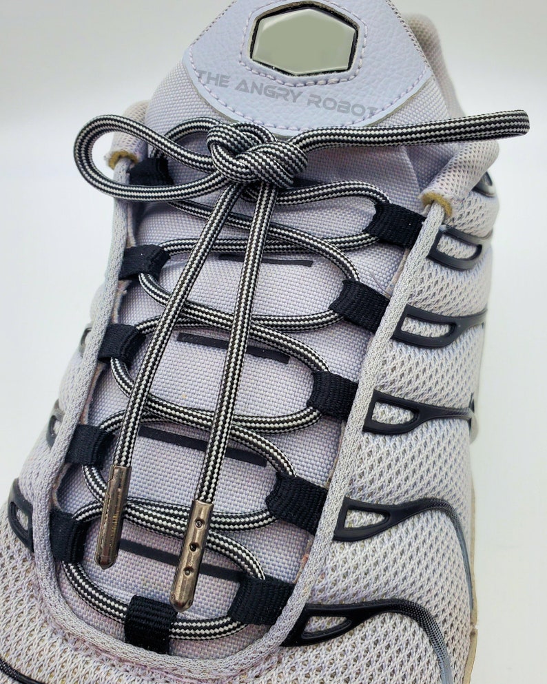 May include: Close-up of a pair of white sneakers with black and white striped shoelaces tied in a knot. The laces are threaded through the eyelets and the text "THE ANGRY ROBOT" is visible on the shoe.