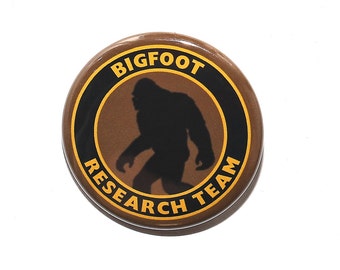 Puede incluir: Un botón marrón con un círculo negro y amarillo alrededor de una silueta de Bigfoot. El texto "BIGFOOT RESEARCH TEAM" está escrito en amarillo alrededor de la silueta.