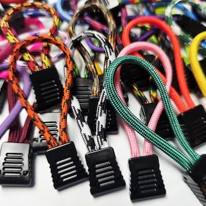 Peut inclure: Une collection de tirettes de fermeture éclair colorées avec des bases en plastique noir. Les tirettes présentent une variété de couleurs de cordon, notamment rose, vert, orange et jaune. Les cordons sont bouclés et fixés à la base de la fermeture éclair.