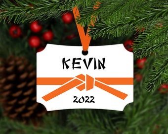 May include: White ceramic Christmas ornament with an orange karate belt, the name "KEVIN" and the year "2022" printed in black.