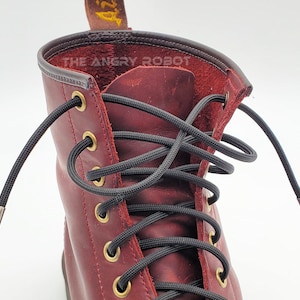 May include: Close-up of a pair of red leather boots with black shoelaces. The laces are threaded through metal eyelets and have metal aglets at the ends. The text "THE ANGRY ROBOT" is visible on the boot.