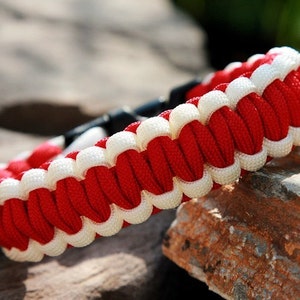 May include: Red and white paracord bracelet with a black buckle closure. The bracelet is made of woven paracord and has a white stripe running along the edge.