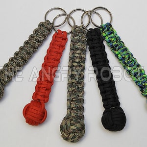 May include: Five different paracord keychains with a knotted ball at the end. The keychains are in various camouflage patterns, including green, black, and red.
