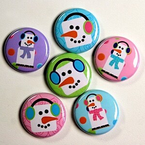 May include: Six colorful round buttons with snowman designs. Each button features a snowman with a different colored background and earmuffs. The snowmen are wearing scarves and have smiling faces.