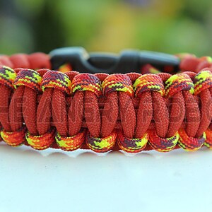Puede incluir: Una pulsera de paracord hecha a mano en tonos de rojo, naranja y amarillo. La pulsera presenta un dise&ntilde;o tejido con un cierre de pl&aacute;stico negro. El texto "THE AN" es visible en la pulsera.