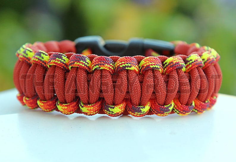 Puede incluir: Una pulsera de paracord hecha a mano en tonos de rojo, naranja y amarillo. La pulsera presenta un dise&ntilde;o tejido con un cierre de pl&aacute;stico negro. El texto "THE AN" es visible en la pulsera.