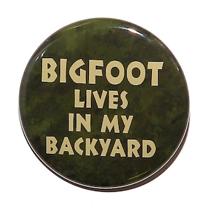 May include: A round button with a green camouflage background and the text "BIGFOOT LIVES IN MY BACKYARD" in gold lettering.