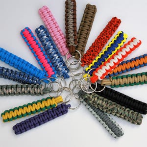 May include: A collection of numerous paracord keychains arranged in a circular fan pattern. Each keychain features a braided cord design in various solid colors and multi-colored patterns, including pink, blue, green, orange, black, and camouflage.