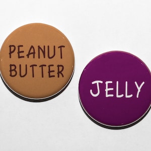 May include: Two round buttons, one brown with the text "Peanut Butter" in brown lettering, and one purple with the text "Jelly" in white lettering.