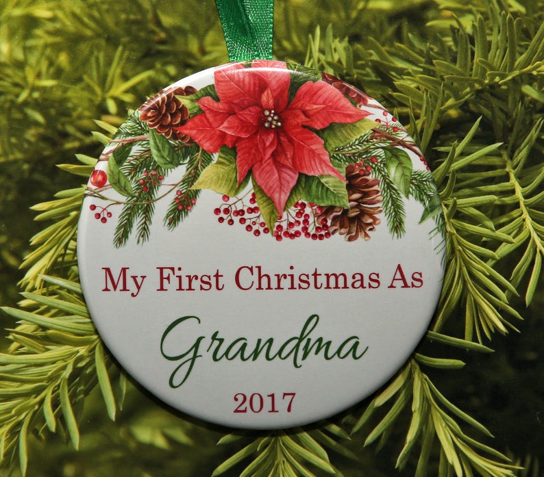 Puede incluir: Adorno navide&ntilde;o redondo blanco con una flor de pascua roja, ramas de pino y pi&ntilde;as. El adorno tiene el texto "My First Christmas As Grandma 2017".