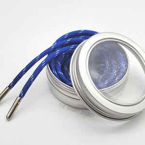 May include: A silver metal container with a clear lid, containing a blue and white shoelace. The container has a silver rim around the lid.