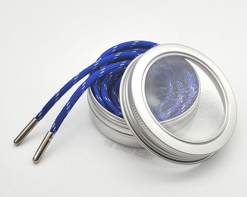 May include: A silver metal container with a clear lid, containing a blue and white shoelace. The container has a silver rim around the lid.