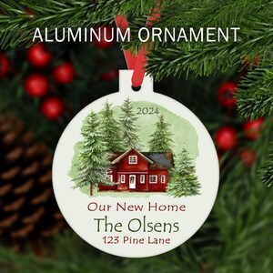May include: A white aluminum Christmas ornament with a watercolor illustration of a red house with green trees. The text "Our New Home The Olsens 123 Pine Lane 2024" is printed on the ornament.