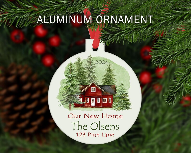 May include: A white aluminum Christmas ornament with a watercolor illustration of a red house with green trees. The text "Our New Home The Olsens 123 Pine Lane 2024" is printed on the ornament.