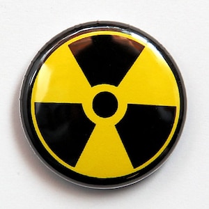 May include: A round, black and yellow radiation symbol pin. The pin features a black border and a yellow trefoil symbol, centered on a black background. The pin is a warning symbol for radioactive materials.