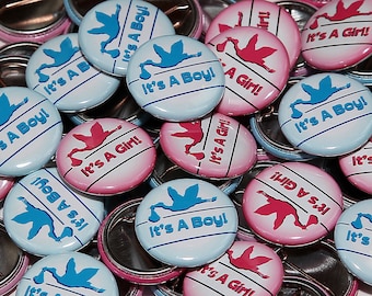May include: A close-up of a pile of baby shower buttons. The buttons are blue and pink and feature a stork carrying a baby. The text on the buttons reads "It's A Boy!" and "It's A Girl!"