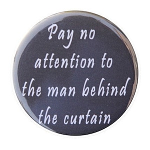May include: A round, black button with white text that reads "Pay no attention to the man behind the curtain."