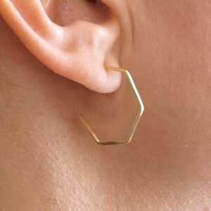 May include: A close-up of an ear with a gold-coloured, geometric hexagon-shaped earring. The earring is a simple, modern design. The skin tone is fair, and the background is blurred.
