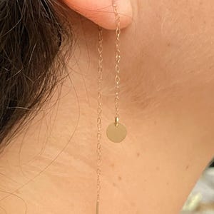 May include: Gold-colored chain earrings. One earring features a small, round disc and a long, thin bar. The other earring has a chain with a small, round disc and a long, thin bar. The earrings are hanging from the ear.