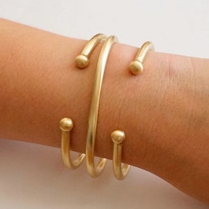 May include: Three gold-toned bangle bracelets with rounded ends. The bracelets are stacked on a person's wrist.