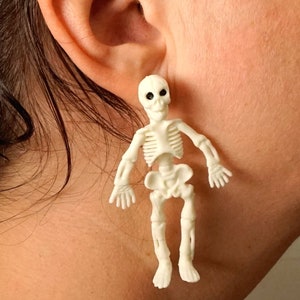 May include: A pair of skeleton earrings. The earrings are white and have black eyes. The earrings are hanging from a person's ear.