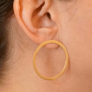 Puede incluir: Un par de pendientes de aro dorados puestos en la oreja de una persona. Los pendientes son simples y de forma circular.