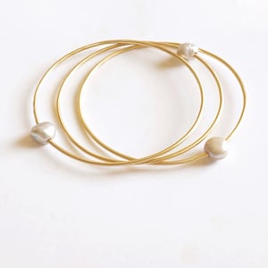 Op de afbeelding: Drie goudkleurige armbanden met zilverkleurige kralen. Eén kraal is hartvormig, de andere twee zijn rond. De armbanden zijn met elkaar verweven en liggen op een witte ondergrond.