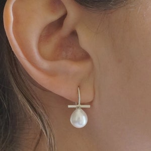 May include: A pair of silver earrings with a small white pearl dangling from each earring. The earrings have a simple, minimalist design.