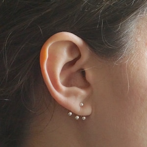 Può includere: Primo piano di un orecchio con tre piccoli orecchini a sfera d'argento. Gli orecchini sono in fila, con i due superiori più vicini tra loro rispetto a quello inferiore.