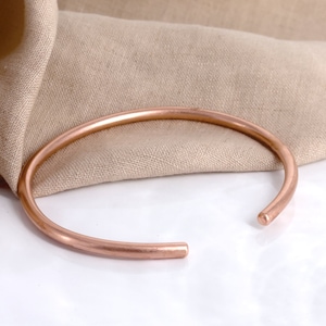 May include: A simple, minimalist rose gold bangle bracelet. The bracelet is made of a single, smooth piece of metal and has a slightly rounded shape.