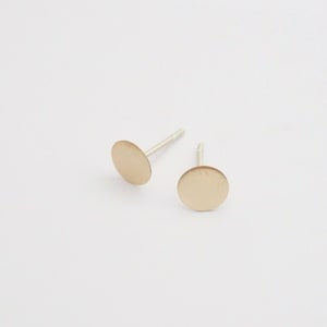 May include: A pair of gold-toned stud earrings with a round, flat design. The earrings are on a white background.