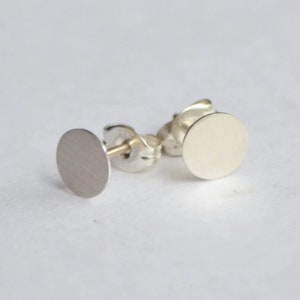 May include: A pair of silver stud earrings with round, flat discs. The earrings are simple and minimalist.
