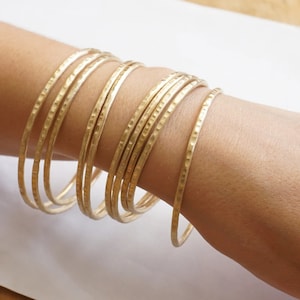 May include: A set of gold-toned hammered metal bangles. The bangles are stacked on a person's wrist.