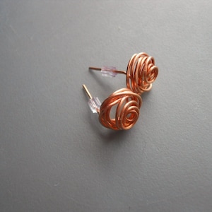Könnte beinhalten: Ein Paar Ohrringe aus Kupferdraht in Rosenform. Die Ohrringe bestehen aus dünnem Kupferdraht, der zu einer Rosenform gedreht und geformt wurde. Die Ohrringe sind an einem Stift mit einer kleinen durchsichtigen Kunststoffrückseite befestigt.