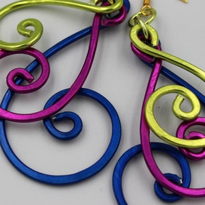 May include: A pair of abstract earrings with a swirling design. The earrings feature a combination of bright colors, including blue, magenta, and yellow-green. The metal earrings have gold-colored hooks.