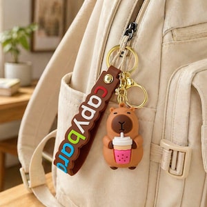 May include: A brown and tan capybara keychain holding a pink and white drink, attached to a brown strap with the word "capybara" in blue and red letters. The keychain is attached to a beige backpack.