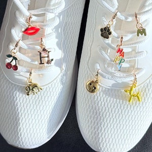 Peut inclure: Paire de chaussures de sport blanches avec des breloques décoratives suspendues aux lacets. Les breloques comprennent une lèvre rouge, une cerise, la lettre "H", un éléphant, une empreinte de patte, la lettre "M", le mot "HAPPY", un smiley et un chien ballon jaune.