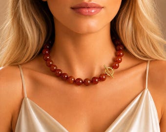 May include: A beaded necklace featuring large, round, reddish-brown stones. The necklace has a gold-colored toggle clasp. The necklace is worn against a cream-colored satin top.