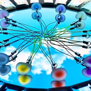 May include: A collection of heart-shaped charms in various colors, including red, blue, yellow, and purple, connected by silver-toned clasps and cords. The charms are arranged in a circular pattern against a reflective surface, with a blue sky background.