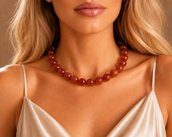 May include: A beaded necklace featuring large, round, reddish-brown beads. The necklace is worn against a background of a cream-colored satin top. The beads appear to be polished and have a glossy finish.