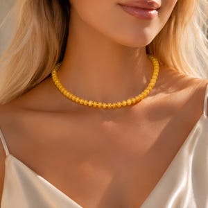 May include: A bright yellow beaded necklace featuring small, uniform, round beads strung together in a single strand. The necklace rests against the neck, showcasing a vibrant, translucent golden-yellow hue that catches the light.